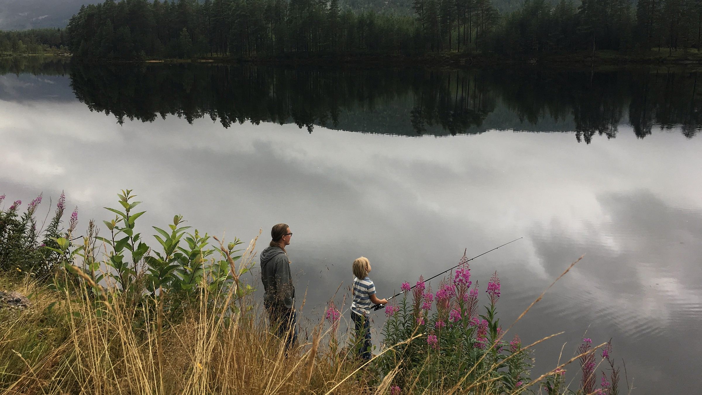 En voksen og et barn står ved bredden av et stille vann, barnet med en fiskestang.